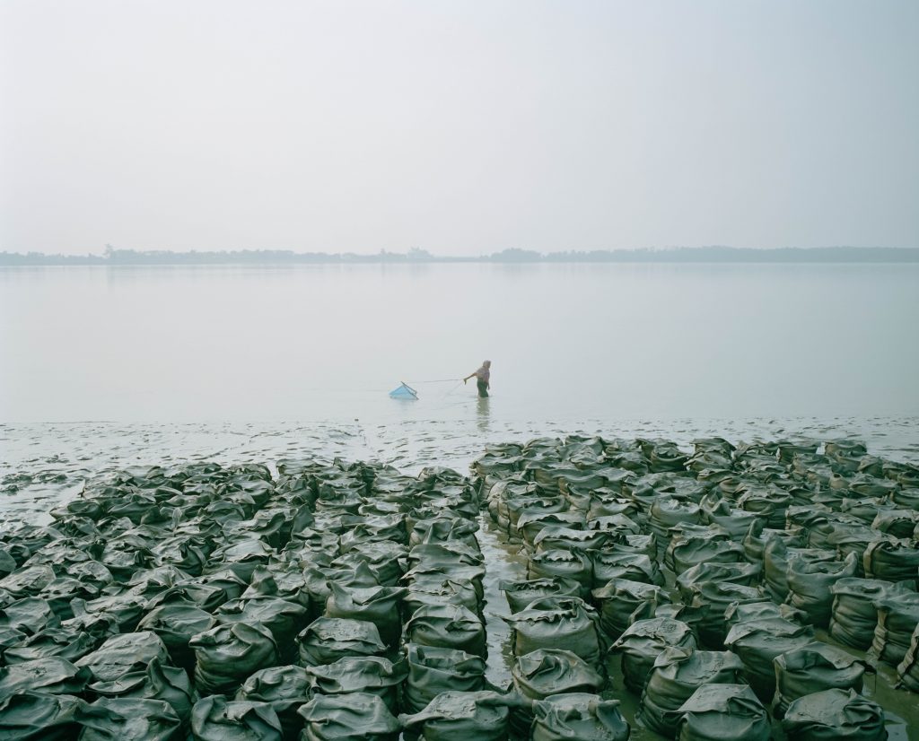 Une femme du Bangladesh marchant dans l'eau, et attrapant de petits poissons ou des crevettes près de sacs de sable trempés dans la rivière. Photographe : Shunta Kimura.