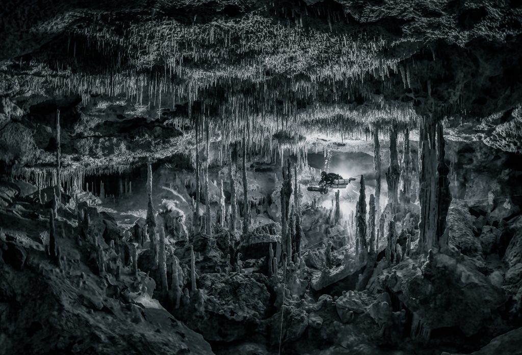 Un plongeur naviguant sur son scooter sous-marin à travers une chambre pleine de stalactites et de stalagmites, montrant la nature brute et les textures étonnantes des grottes inondées au Mexique. Photo : Martin Broen.